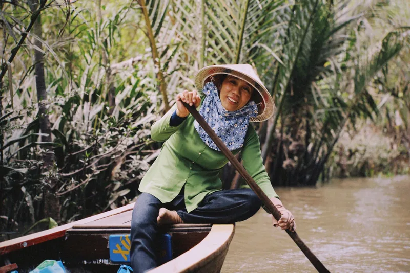 A woman in a conical hat rows a wooden boat through a palm-lined canal in the Mekong Delta.
