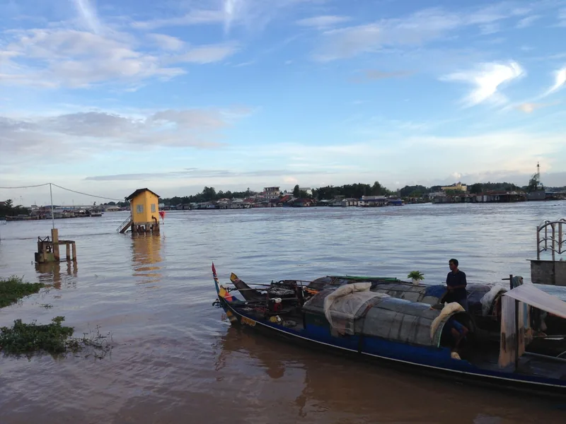A loaded wooden boat with a man aboard rests on the wide Chau Doc river under a stilt house and evening sky.