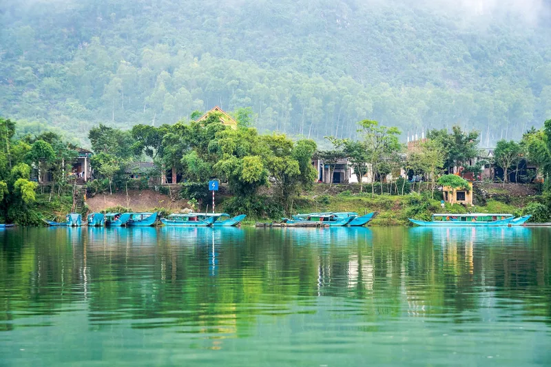 Blue tour boats line the riverbank at Phong Nha with misty limestone mountains rising behind a small village.