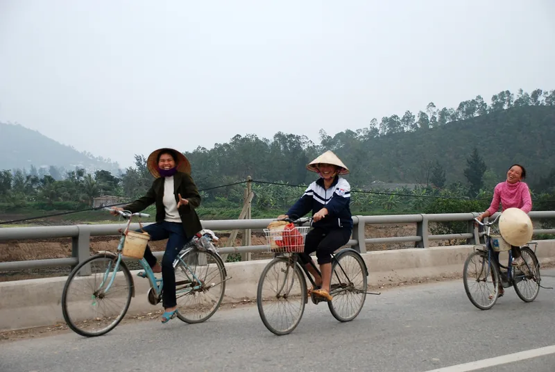 Three women in conical hats ride bicycles across a rural bridge near Hanoi, smiling at the camera.