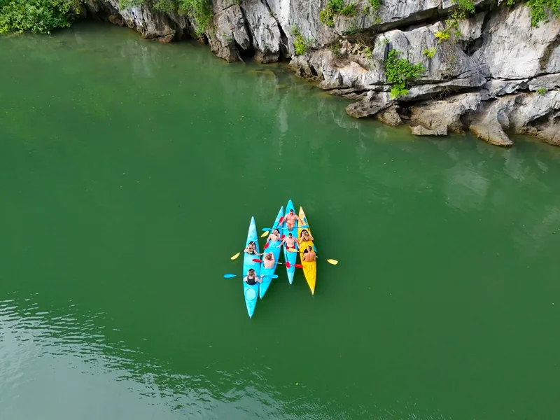 Kayakers paddle colorful boats across emerald water beneath limestone cliffs in Lan Ha Bay.
