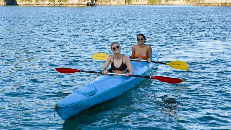 Two women in swimsuits paddle a blue kayak on calm turquoise water near limestone islands in Lan Ha Bay.
