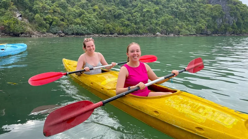 Two women paddle a yellow kayak on calm green water beneath limestone karsts in Lan Ha Bay.