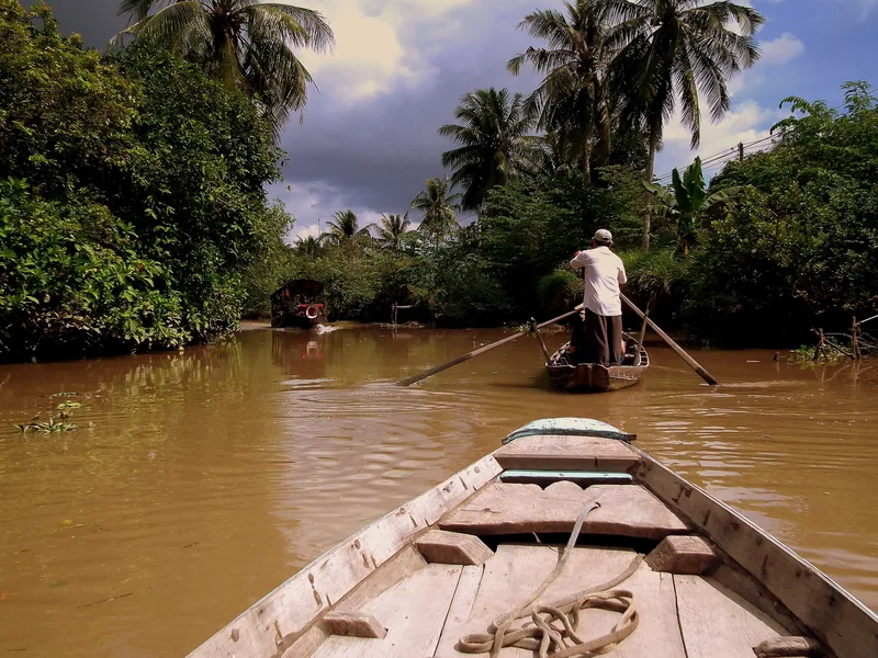 A boatman rows a wooden boat along a muddy, palm-lined canal in the Mekong Delta, Vietnam.