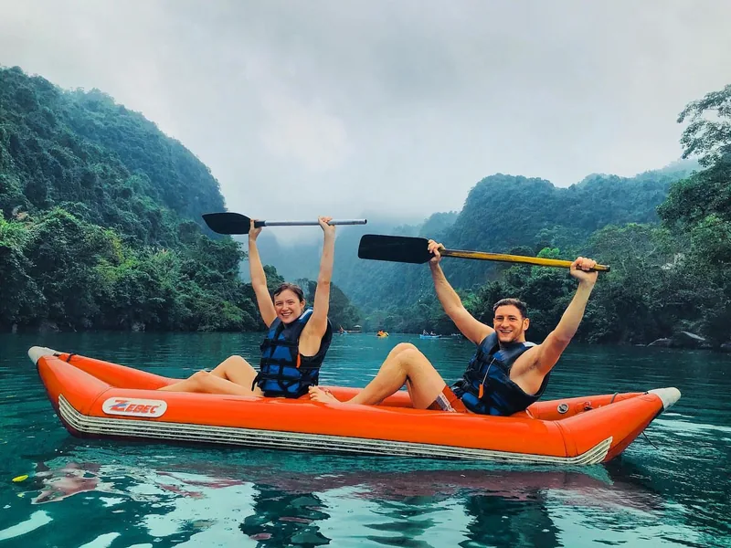 A couple raises paddles while kayaking on emerald water surrounded by misty limestone karsts near Song Chay river, Quang.