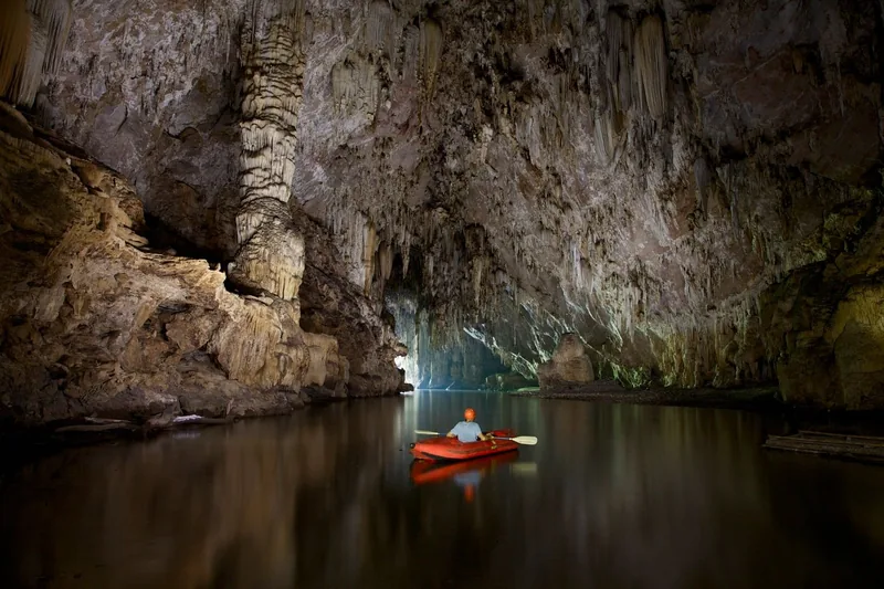 A lone kayaker paddles a red raft through the still, dark waters of Phong Nha Cave beneath towering limestone formations.