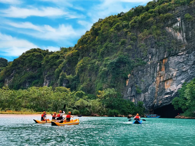 Kayakers in life vests paddle turquoise water toward a limestone cave entrance at Phong Nha, surrounded by forested karst.