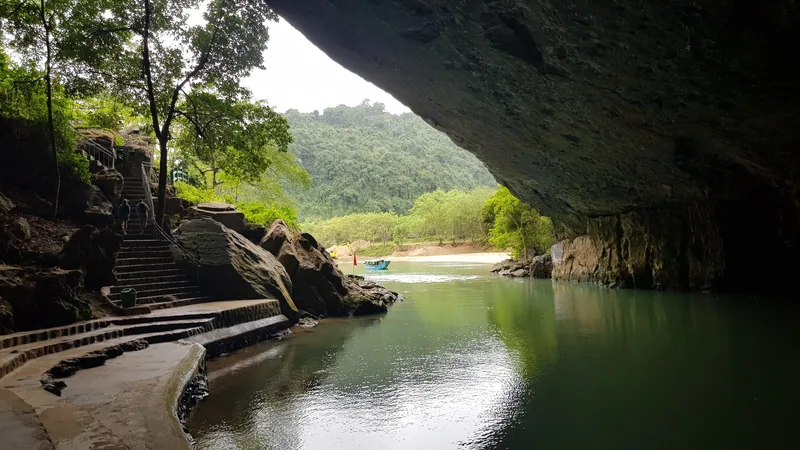 A stone stairway leads beside a calm river flowing beneath a cave entrance surrounded by jungle in Phong Nha.