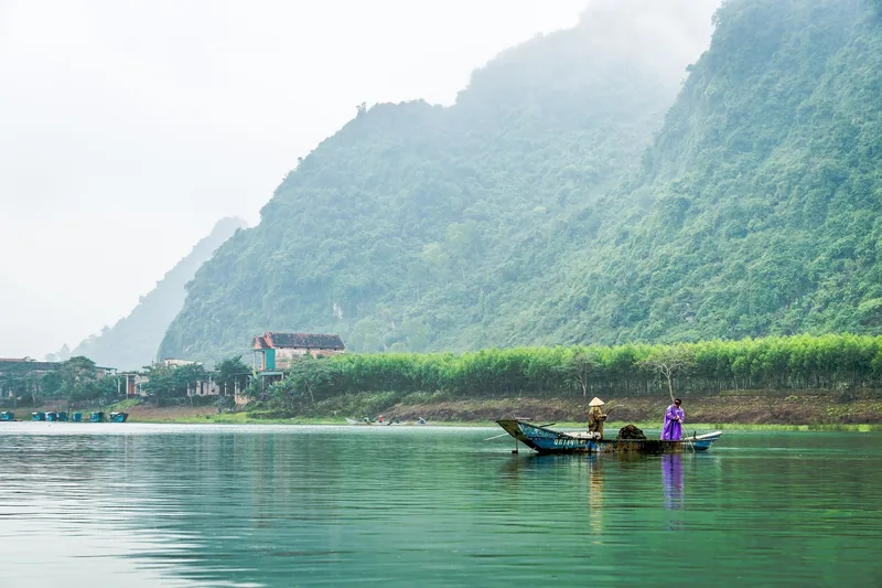 A woman in a purple dress stands on a small boat on the river at Phong Nha, framed by misty limestone mountains.
