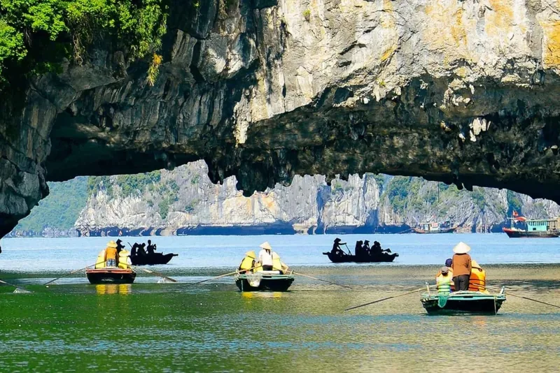 Small rowboats carrying tourists pass beneath a rocky stone arch on a limestone-fringed bay in Vietnam.