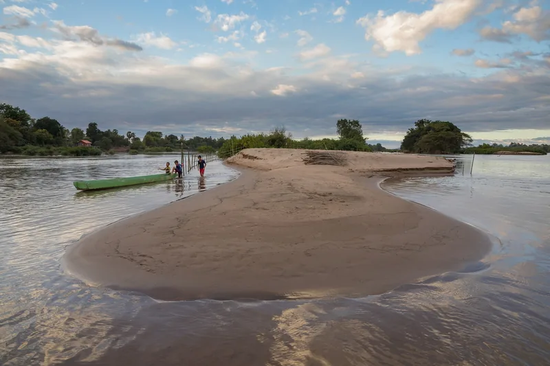 A sandy river island with wading children and a green boat sits amid the Mekong's braided channels at Si Phan Don.