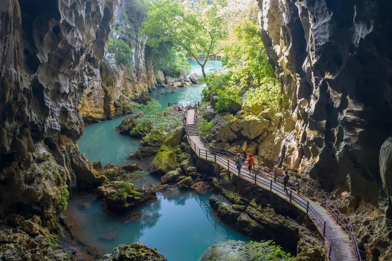 A wooden walkway winds along a turquoise river through limestone karst walls near Quang Binh's caves.